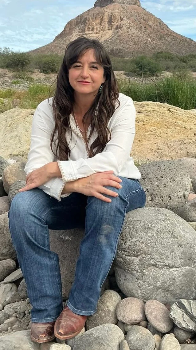 Carla Puentes seated on river rocks in Baja, with a rust-colored mesa behind her